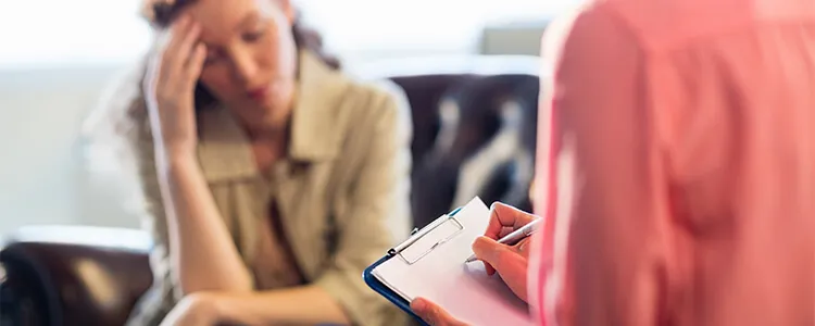 A woman with her head in her hand talks to a clinician, who takes notes on a clipboard.